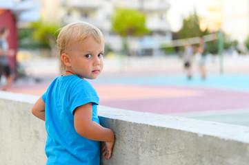 Naklejka premium a little three year old boy in a blue t-shirt watches teenagers playing with a ball on an outdoor sports field, dreaming of playing sports