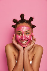 Vertical shot of natural beauty model touches face gently, smiles sincerely, wears cosmetic mask to avoid wrinkles and blackheads, dresses bath towel around body, isolated on pink background