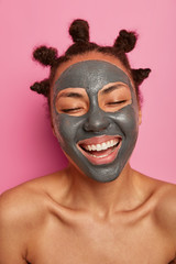 Happy ethnic woman enjoys applying face mask, purifying skin, does skin care anti aging beauty treatment, has bun hairstyle, naked body, laughs positively with closed eyes, broad toothy smile