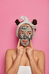 Asian young woman points at face with applied beauty mask, has everyday morning routine, feels happy, cares about skin, has two hair buns, stands wrapped in soft towel, isolated on pink background