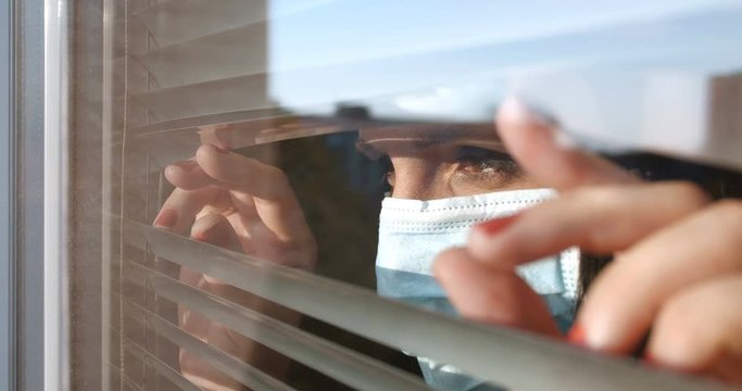Close up profile portrait from indoors of woman wearing medical face shield pushing blinds at window with hands. Looking fearfully outside being afraid of germs. Concept of quarantine, self-isolation