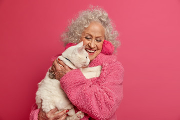 Old people stay at home during quarantine. Happy elderly curly woman dressed in domestic robe, holds white cat on hands, being on self isolation, feels not lonely in company of favorite pet.