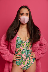 Displeased Asian woman wears sterile mask, protects herself from coronavirus or flu, wears swimsuit and domestic robe, stands against rosy background, has respiratory disease, being at isolation