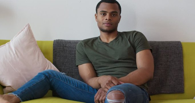 Portrait of attractive black millennial man sitting on couch looking at camera. Medium wide shot of African American homeowner in his living room. 4k slow motion handheld
