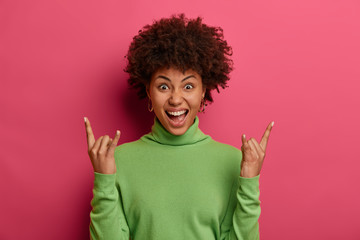 Sassy cool ethnic woman acts cheeky, makes rock n roll gesture, heavy metal sign, exclaims loudly, enjoys favorite song, has fun at concert, enjoys party, wears green turtleneck, isolated on pink wall