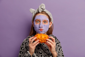 Little redhead adorable girl applies purple face mask, has regular beauty treatments, holds horned melon, looks directly at camera, blows cheeks, wears bathrobe, isolated on violet background.