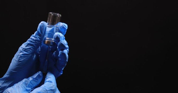 Close-up macro shot of hands in latex blue medical gloves holding an ampoule with clear liquid and collecting it with syringe. On black studio background isolated with free space for inscription.
