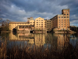 Old abandoned factory in the italian countryside, along a river, reflected on water, under a cloudy sky.