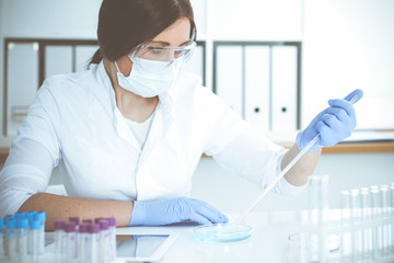 Professional female scientist in protective eyeglasses and mask making experiment with reagents in...