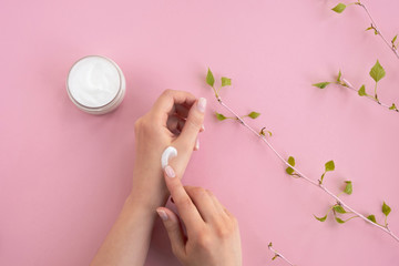 Cosmetic cream on woman's hands. Jar with cream and a branch with green leaves on light pink background. Beauty concept, hand skin care. Top view, flat lay