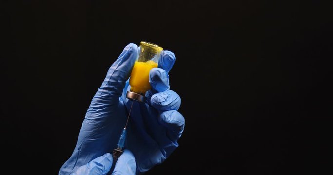 Close-up macro shot of hands in latex blue medical gloves holding ampoule with yellow liquid and collecting it with syringe. On black studio background isolated with free space for inscription.
