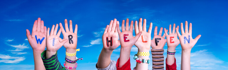 Kids Hands Holding Colorful German Word Wir Helfen Means We Help. Blue Sky As Background