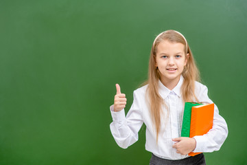 Smart girl stands with books near a blackboard and shows thumbs up gesture. Empty space for text