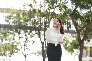 Smiling woman calling on phone outdoors. Pretty young lady standing with city view in background. City walk concept. Side view.