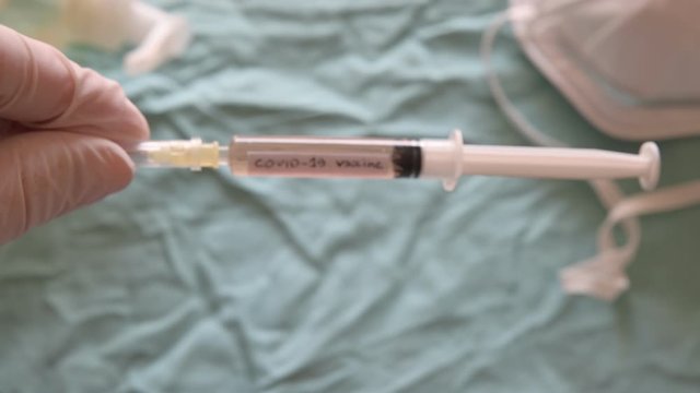 Blurred image of the sanitizing gel, a mask and a syringe. Gloved hand raise the syringe in a close-up image. It reads coronavirus vaccine.