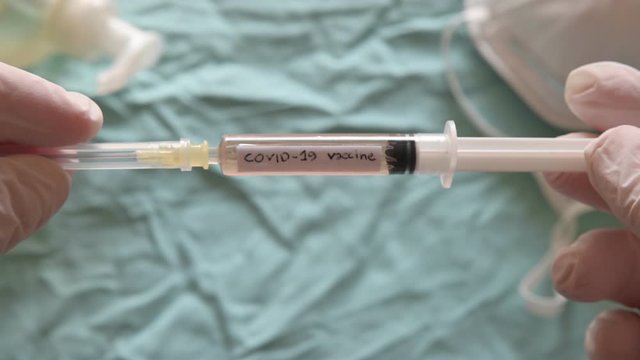 Blurred image of the sanitizing gel, a mask and a syringe. Gloved hands raise the syringe in a close-up image. It reads coronavirus vaccine.