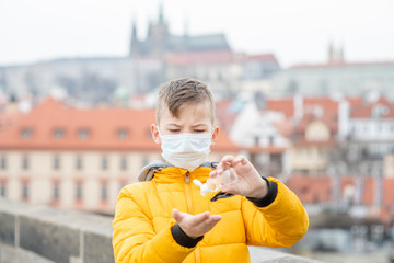 Boy wearing medical mask uses sanitizer to disinfect hands on the background of Prague Castle....