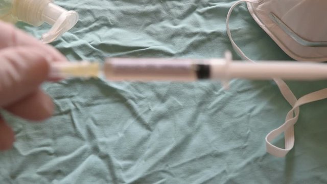 Blurred image of the sanitizing gel, a mask and a syringe. Gloved hands raise the syringe in a close-up image. It reads coronavirus vaccine.