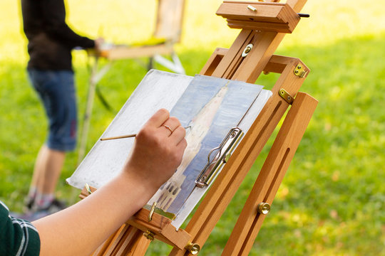 artist painting on open air, wooden easel sketchbook with unfinished painting close-up. Female hand paints in a sunny summer meadow