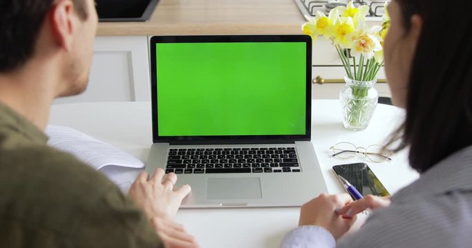 Young husband and wife in kitchen video chatting with their friends or family using laptop MacBook Pro with green screen. Millennial White man and Caucasian woman facetime with another couple.