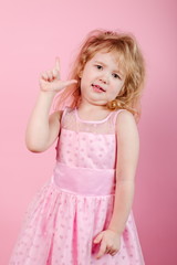 Little funny girl in a pink dress standing on a pink background showing thumb up