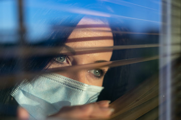 Woman with surgical mask looking out of home window, concept of quarantine during viral pandemic...