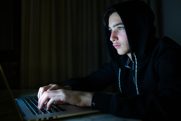 Hacker is  hacking into the computer network. Young boy in a dark room with laptop.  Hacker with a...
