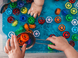 Toddlers sort by color the colored caps that they take out of the water. The development of children's motor skills using a sensory box. Development of children at home during a quarantine - obrazy, fototapety, plakaty