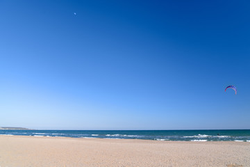 Sea shore with a sandy beach, a sunny day shows the moon and a lone kite surfer.