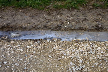 rural scene with frozen water puddle between car tracks in wet soil and shingle stones, puddle on dirt road in late winter