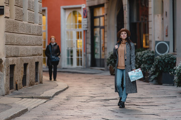 A young girl tourist in a fashionable brown hat is walking along a small street with cobblestones. The girl is going our way. Against the background of beautiful green plants.