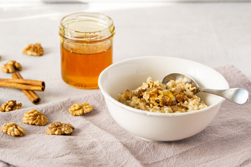 Oatmeal porridge with walnuts, cinnamon, and honey in a bowl on grey napkin. There is jar of honey, walnuts and cinnamon stick. Healthy vegetarian breakfast.