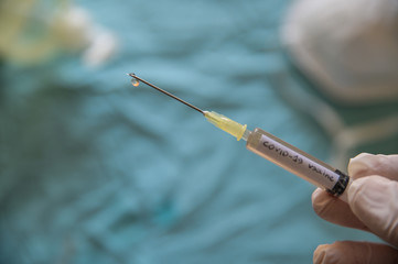 Close up shot of a syringe with the word coronavirus vaccine.The hand holding it is pressing the plunger and a drop is coming out of the needle. In the blurred background a mask and the sanitizing gel