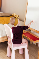 Little girl draws with felt-tip pen on a blackboard in a bright and stylish children's room. Baby sitting on a chair 