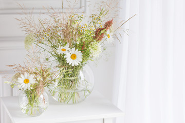 Bouquet of gentle camomile in white cup. Morning light in the room. Soft home decor,  vase with white flowers on  white wall background and on wooden table. Interior. Greeting card. Copy space.