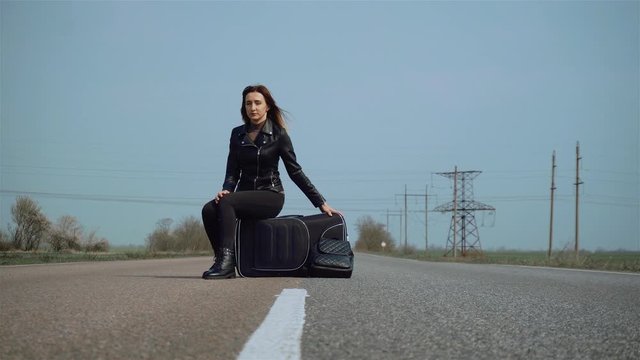 Woman tourist sit on her luggage in suitcase on wheels in middle of asphalt road and waiting for passing car. Female hitchhiking around country