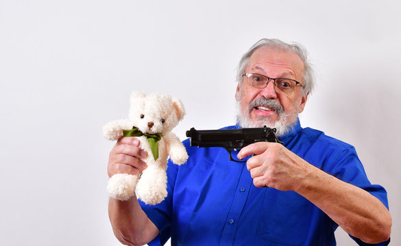 Old man in blue shirt and bow tie threatens to shoot a white teddy bear with a 9mm automatic pistol.