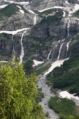 Obraz premium A multi-jet alpine waterfall on a high cliff with a green forest at the foot with glimpses of snow. The concept of a natural landscape in a natural environment on a cloudy day.