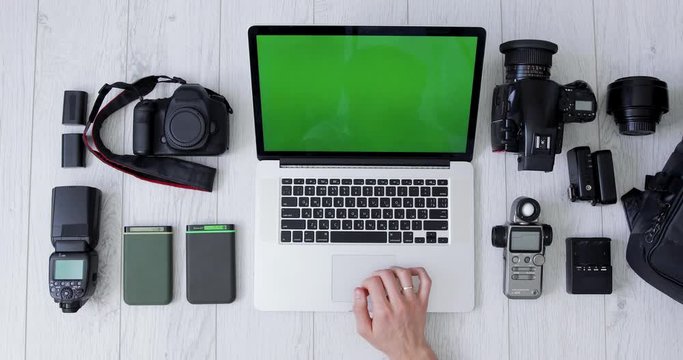 Top View of a Photographer Hand Processing Photos on His Laptop With Green Screen. Backpack, Medium format film and DSLR Camera, Lens, External Hard Drives and Light Meter Lie Beside of Him