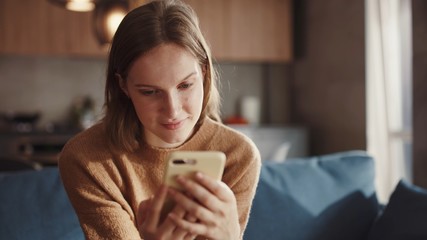 Charming goldhair teenager girl sitting of the sofa at home using phone and smiling. Feeling happy. Communication female looking message at cell phone or smartphone