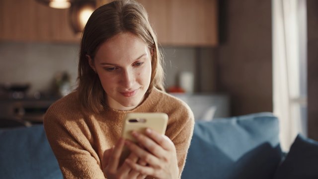 Charming goldhair teenager girl sitting of the sofa at home using phone and smiling. Feeling happy. Communication female looking message at cell phone or smartphone