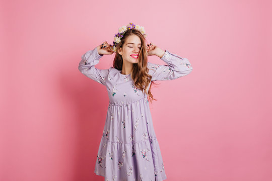 Blissful Girl In Flower Wreath Posing With Eyes Closed. Indoor Portrait Of Happy Caucasian Female Model In Retro Purple Dress Enjoying Photoshoot.