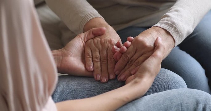 Close up young woman holding wrinkled hands of elderly retired mother, showing support care love. Two female family generations enjoying trustful conversation, sitting on comfortable sofa at home.