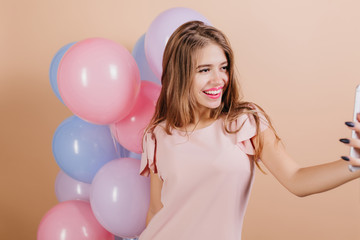 Fototapeta na wymiar Enchanting birthday girl with bright lipstick making selfie with balloons. Studio shot of smiling charming young woman with wavy hair having fun during party.