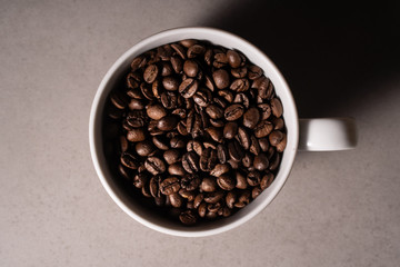 Zdjęcie białego kubka z ziarnami kawy na szarym tle, dookoła rozsypane ziarna kawy