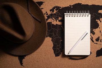 Zdjęcie kapelusza i notesu z długopisem na tle mapy świata - obrazy, fototapety, plakaty