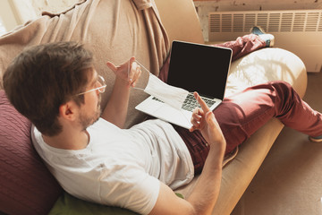 Young man studying at home during online courses for disinfector, nurse, medical services. Getting profession while isolated, quarantine against coronavirus. Using laptop, smartphone, headphones.