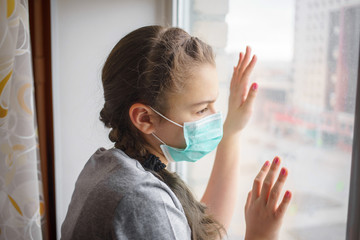 a girl in a medical mask looking through window. at the quarantine caused by coronavirus