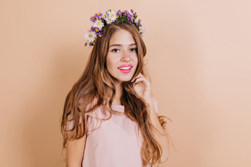 Dreamy long-haired brunette woman with purple flowers in hair smiling to camera. Interested pretty girl standing on light background.