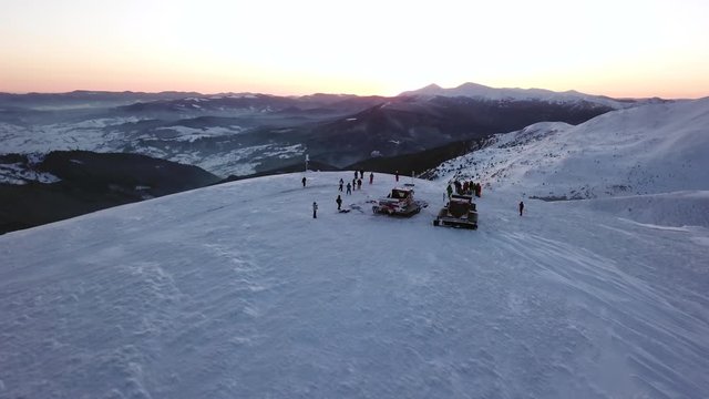 People stand on the edge of the mountain at sunrise, aerial shot. Dawn on the mountain, people stand about two snowcats and watch the sun rise. Aerial shot fly away.